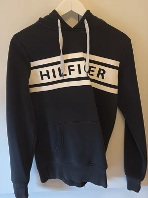 Svart hoodie Tommy Hilfiger S - Snygg svart hoodie från Tommy Hilfiger med vit logga och breda vita ränder över bröstet. Klassisk huva med vita snören och stor magficka framtill. Tillverkad i mjukt bomullsmaterial som känns skönt mot huden. Perfekt för en avslappnad och trendig look.