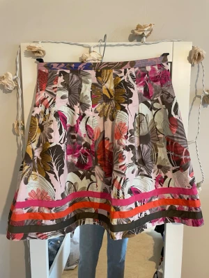 Blommig kortkjol med färgglada ränder - Säljer en kortkjol med blommigt mönster i rosa, rött, brunt och gult. Kjolen har färgglada ränder i rosa, orange och brunt längst ner. Perfekt för dig som vill sticka ut med en unik och färgsprakande stil.