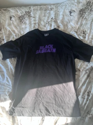 Svart Black Sabbath t-shirt H&M - Säljer en svart t-shirt från H&M med lila 'Black Sabbath'-tryck på bröstet. Klassisk rund hals och korta ärmar. Perfekt för dig som gillar bandtröjor och vill ha en enkel men snygg look.