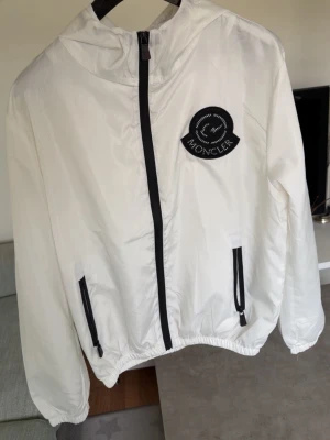 Vit vindjacka från Moncler - Snygg vit vindjacka från Moncler med svart dragkedja och svarta detaljer på fickorna. Stor Moncler-logga på bröstet och mindre logga på ärmen. Jackan har huva och elastiska muddar vid ärmslut och midja. Perfekt för blåsiga dagar.