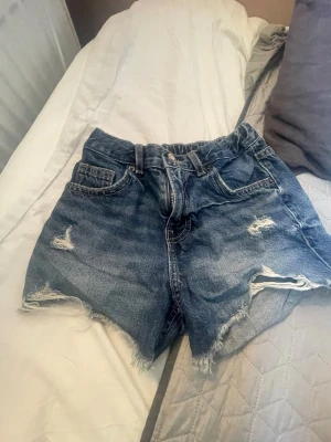 Blåa högmidjade jeansshorts - Säljer ett par blåa högmidjade jeansshorts med slitna detaljer och fransig kant. Klassisk femficksmodell med dragkedja och knapp framtill. Perfekta för sommaren och ger en avslappnad vibe.