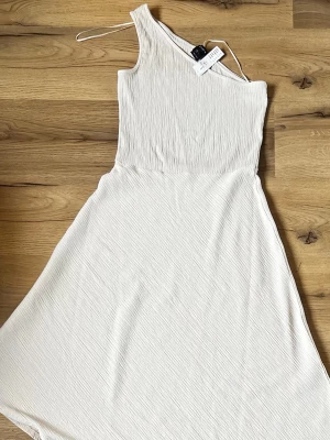 Off white oneshoulder klänning från Lindex - Säljer en stilren vit oneshoulder klänning från Lindex. Klänningen har en asymmetrisk axel med smal axelrem och är tillverkad i ett lätt, strukturerat tyg som ger en snygg effekt. Aldrig använd original pris är 399