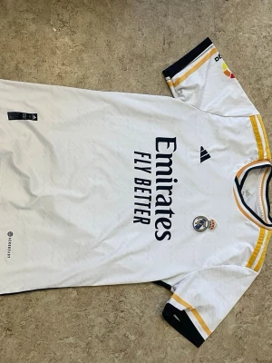 Real Madrid matchtröja Vini Jr 20 - Säljer en vit Real Madrid matchtröja från Adidas med Vini Jr och nummer 20 på ryggen. Tröjan har gula och svarta detaljer på ärmarna, klubbmärke och sponsortryck på bröstet samt La Liga-märke på ärmen. Tillverkad i lätt och ventilerande material.