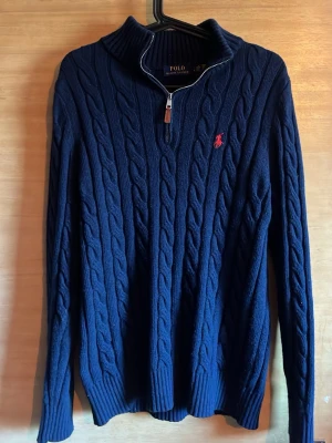 Mörkblå kabelstickad tröja Ralph Lauren - Mörkblå kabelstickad tröja från Polo Ralph Lauren med halv dragkedja och hög krage. Tröjan har ett rött broderat logomärke på bröstet och är gjord i ett mjukt, tjockt material. Perfekt för lager på lager och en klassisk look. Sitter ungefär som M. Fråga om längd osv 