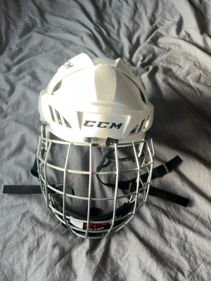 Vit hockeyhjälm från CCM - Säljer en vit hockeyhjälm från CCM med galler i metall och justerbara remmar. Hjälmen har svart vaddering på insidan för extra komfort och skydd. Perfekt för dig som spelar ishockey och vill ha bra säkerhet på isen.