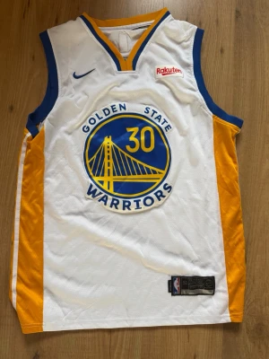 Golden State Warriors baskettröja Steph Curry - Golden State Warriors basketlinne i vitt med blå och gula detaljer. Nummer 30 och Curry på ryggen. Stort Warriors-logotyp på framsidan och Rakuten-logga. Tillverkat i lätt och ventilerande meshmaterial, perfekt för basket eller träning. Pris kan diskuteras
