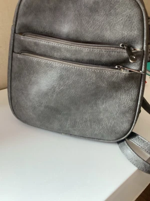 Grå fyrkantig ryggsäck i konstläder - Stilren grå ryggsäck och handväska i konstläder med två framfickor med dragkedja och justerbara axelband. Väskan har en fyrkantig form och en enkel 3 stora fickor , modern look som passar till det mesta. Perfekt för dig som vill ha en arbetsmaterial (titta på sista bilden)