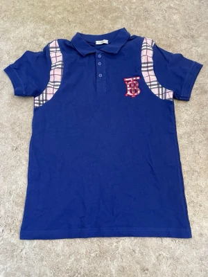 Blå pikétopp med rutiga detaljer - Snygg blå pikétopp från Burberry med klassiska rutiga paneler i beige, svart och rosa på axlarna. Toppen har korta ärmar, krage med knappar och ett rött broderat TB-märke på bröstet. Perfekt för en trendig och avslappnad stil.