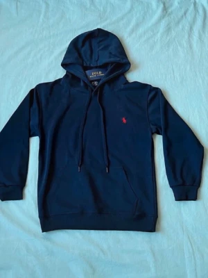 Mörkblå hoodie från Polo Ralph Lauren - Aldrig använd!!!Mörkblå hoodie från Polo Ralph Lauren med klassisk huva och dragsko. Tröjan har en liten röd broderad logga på bröstet och en känguruficka framtill. Tillverkad i mjukt bomullsmaterial och har ribbade muddar vid ärmslut och nederkant.