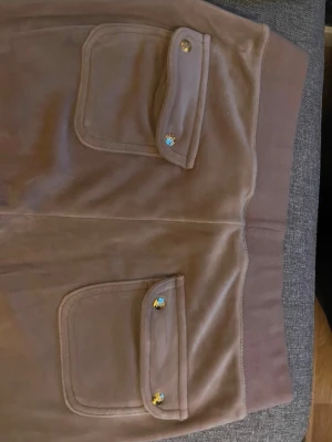 Beiga mjukisbyxor med fickor - Säljer ett par beiga mjukisbyxor i mjukt velourmaterial. Byxorna har två stora fickor bak med lock och guldfärgade knappar. Resår i midjan för skön passform och långa ben. Perfekta för chill dagar eller när du vill vara extra bekväm.