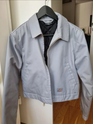 Ljusgrå bomberjacka från Dickies - Snygg ljusgrå bomberjacka från Dickies med klassisk krage, dragkedja framtill och två sidofickor. Jackan har quiltat svart innerfoder och är tillverkad i ett slitstarkt bomullsmaterial. Kortare modell med justerbar knappslå bak. Perfekt för en streetwear-look.