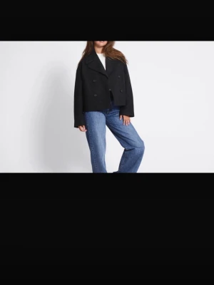 Svart dubbelknäppt ullrock - Stilren svart rock i ullblandning med dubbelknäppning och bred krage. Kortare modell som ger en modern look och passar perfekt till jeans eller byxor. Klassisk design utan synliga loggor eller detaljer.