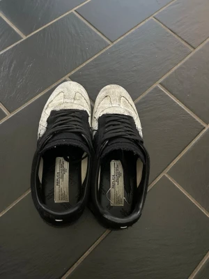 Svarta och vita sneakers från Maison Margiela - Unika sneakers från Maison Margiela i svart och vitt med coolt, slitet färgmönster. Skorna har lågt skaft, svarta snören och klassisk gummisula med tydlig struktur undertill. Perfekta för dig som gillar statement-plagg och streetwear.