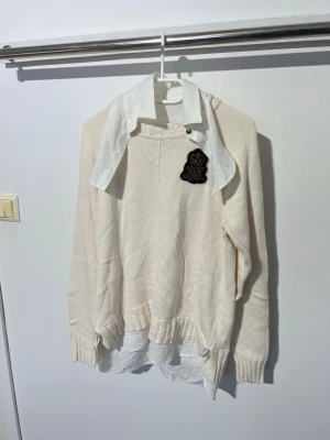 Beige stickad tröja med skjortkrage Lauren Ralph Lauren - Säljer en beige stickad tröja med vit skjortkrage och dekorativt svart märke på bröstet. Tröjan har långa ärmar, ribbade muddar och en lös passform. Perfekt för en trendig lager-på-lager-look.