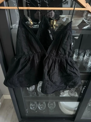 Svart broderad linnetopp från Zara - Säljer en svart linnetopp från Zara med tunna axelband och v-ringad front. Toppen har broderade detaljer och spets längs kanterna för en extra snygg touch. Modellen är croppad och har en luftig, voluminös passform som känns både trendig och bekväm.