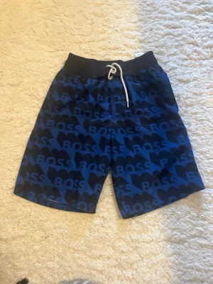 Blå badbyxor från BOSS - Snygga blå badbyxor från BOSS med ett stort logomönster över hela plagget. Storlekn är 9-10 år 