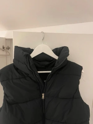 Svart dunväst med hög krage - Snygg svart dunväst med hög krage och dragkedja framtill. Västen har en quiltad design och är perfekt för lager-på-lager-stil. Tillverkad i ett mjukt och vadderat material som håller värmen. Enkel och stilren look som passar till det mesta.