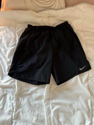 Svarta träningsshorts från Nike - Säljer ett par svarta träningsshorts från Nike med elastisk midja och klassisk Nike-logga på benet. Shortsen har en loose passform och är tillverkade i ett lätt syntetmaterial som passar perfekt för sport och aktivitet. Storlek S