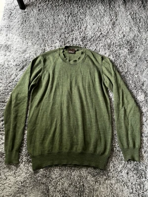 Olivgrön stickad tröja Oscar Jacobson - Snygg olivgrön stickad tröja från Oscar Jacobson i tunn ullmix. Tröjan har rund halsringning, långa ärmar och ribbade muddar vid ärmslut och nederkant. Perfekt för lager på lager och stilrena outfits.
