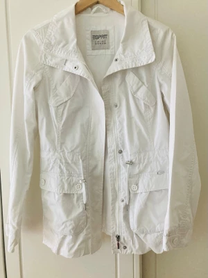 Vit vindjacka från Esprit - Stilren vit vindjacka från Esprit med hög krage, dragkedja och knappar framtill. Jackan har fyra fickor med lock och knapp, justerbar midja och långa ärmar. Tillverkad i lätt material som passar perfekt för blåsiga dagar.