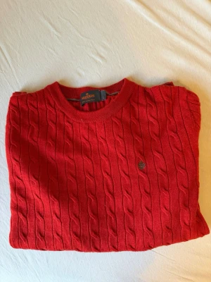 Röd kabelstickad tröja från Morris - Snygg röd kabelstickad tröja från Morris med rund halsringning och diskret logga på bröstet. Tröjan har långa ärmar och ribbade muddar vid ärmslut och nederkant. Perfekt för dig som gillar klassisk stil med en modern twist. Tröjan passar för både M och L