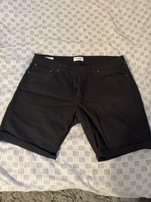 Svarta jeansshorts från Jack & Jones - Snygga svarta jeansshorts från Jack & Jones med klassisk femficksmodell och uppvikta benslut. Shortsen har knappgylf och bälteshällor, perfekta för en avslappnad och stilren look. Materialet är slitstarkt denim i bomull.
