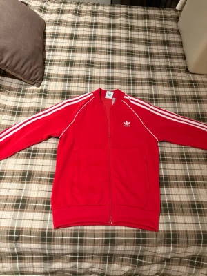 Röd Adidas hoodie med vita ränder - Säljer en röd hoodie från Adidas med klassiska vita ränder längs ärmarna och Adidas-logga på bröstet. Dragkedja framtill och två fickor. Sportig och stilren design, perfekt för en avslappnad look.