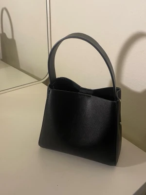 Svart handväska från Gina Tricot  - Säljer en svart väska från Gina Tricot i bra skick. Väskan är sparsamt använd och har ett längre axelband som kan justeras till önskad längd med hjälp av banden på sidorna. Den har en praktisk storlek som rymmer det viktigaste samt ett innerfack med dragkedja för värdesaker. Passar perfekt till både vardag och fest.