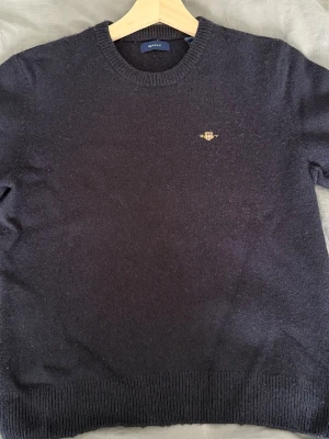 Mörkblå stickad tröja från Gant - Snygg mörkblå stickad tröja från Gant med rund halsringning och diskret broderad logga i gult på bröstet. Tröjan har ribbade muddar vid ärmslut och nederkant. Perfekt för dig som gillar klassisk och stilren design.