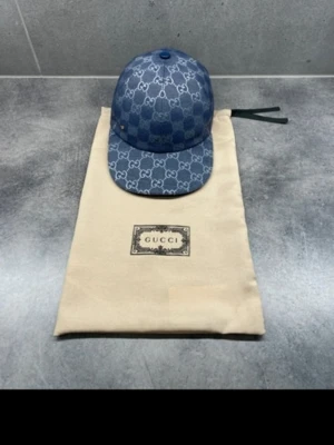 Blå Gucci keps med monogram - Säljer en blå keps från Gucci med det klassiska GG-monogrammet i vitt över hela kepsen. Kepsen har böjd skärm och justerbar rem bak. Materialet ser ut att vara denim eller canvas, vilket ger en cool och trendig look. Perfekt accessoar för dig som gillar streetwear och designer. BOX INGÅR 