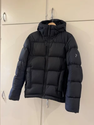 Svart pufferjacka med huva från Peak Performance - Säljer en svart pufferjacka från Peak Performance med huva och dragkedja framtill. Jackan har två stora fickor på sidorna och diskret logga på ärmen. Perfekt för kalla vinterdagar tack vare den tjocka vadderingen och den klassiska boxiga passformen.   Nypris 4200kr