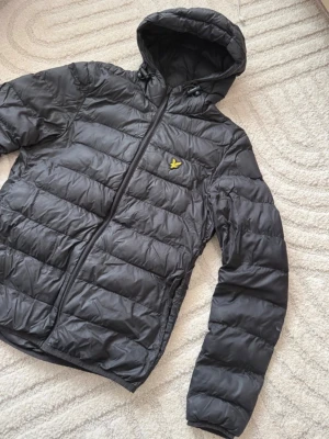 Svart pufferjacka från Lyle & Scott - Svart pufferjacka från Lyle & Scott med huva och dragkedja. Jackan har ett quiltat mönster och den klassiska gula logon på bröstet. Perfekt för kalla dagar och tillverkad i syntetmaterial som håller värmen. Betalning via vanlig swish! 