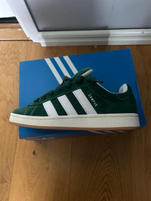 Adidas Campus gröna sneakers - Gröna Adidas Campus sneakers i mocka med klassiska vita ränder på sidorna och vit sula. Skorna har lågt skaft och vita detaljer på häl och plös. Snygg retrostil och tidlös design som passar till det mesta. Levereras i originalkartong.