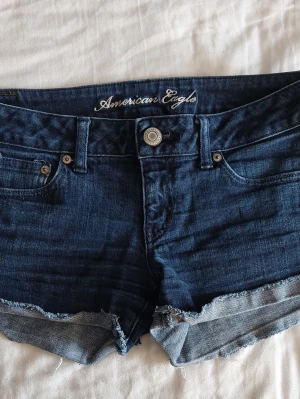 Mörkblå jeansshorts American Eagle - Passar S-M. Snygga mörkblå jeansshorts från American Eagle med råa kanter och klassisk femficksdesign. Shortsen har knapp och dragkedja framtill samt bälteshällor. Perfekta för varma dagar och ger en avslappnad look.