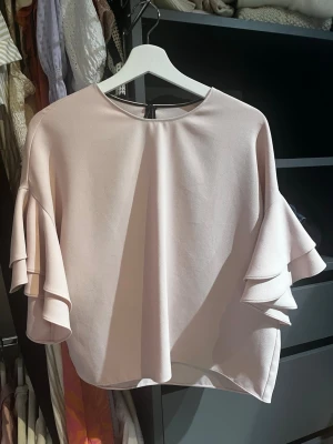 Ljusrosa blus med volangärmar - Supersöt ljusrosa blus med rund halsringning och volangdetaljer på ärmarna. Blusen har en lös passform och är tillverkad i ett mjukt, vävt material som ger ett elegant fall. Perfekt för dig som gillar en feminin och trendig stil.