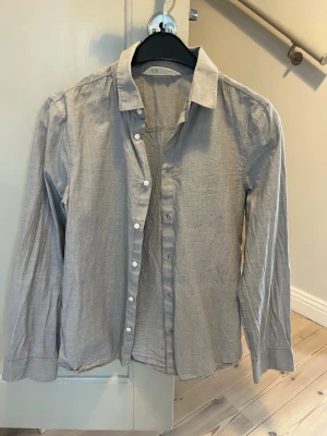 Ljusgrå skjorta från H&M - Klassisk ljusgrå skjorta från H&M med lång ärm och vita knappar framtill. Skjortan har en avslappnad passform och är tillverkad i mjuk bomull
