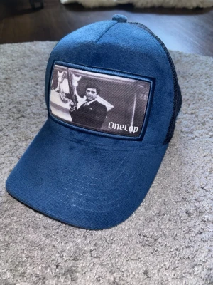 Blå truckerkeps från OneCap - Snygg blå truckerkeps från OneCap med svart mesh på baksidan och ett stort tygmärke framtill med svartvit motiv. Kepsen har böjd skärm och justerbar snapback. Perfekt för dig som vill ha en unik och trendig accessoar.