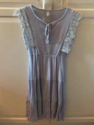 Ljuslila klänning med spetsdetaljer - Söt ljuslila klänning med volang och virkade spetsdetaljer på axlarna. Klänningen har knyt framtill vid halsen och är tillverkad i ett tunt och skönt material. Den har en avslappnad passform och faller fint. Köpt i Sydkorea. Storlek One Size, men passar S. I fint skick. Färgen är syrenlila typ.