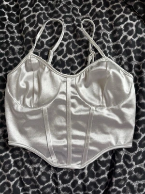 Vit satin korsettinspirerad topp - Snygg vit korsett i glansigt satinmaterial med smala justerbara axelband och markerade sömmar framtill. Mjukt material. Perfekt för att skapa en trendig och elegant look. Aldrig använd. Tvättad 1 gång. 