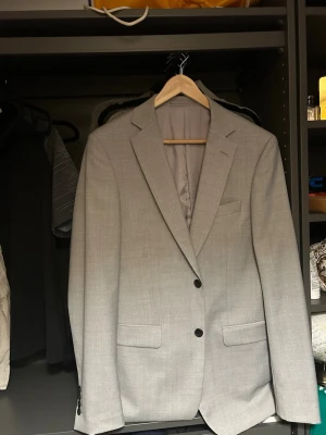 Ljusgrå klassisk kavaj herr - Snygg ljusgrå kavaj med två knappar framtill och fyra dekorativa knappar vid ärmslutet. Kavajen har två fickor med lock och en bröstficka. Tidlös modell med smal passform och helfodrad insida. Perfekt för en stilren look.