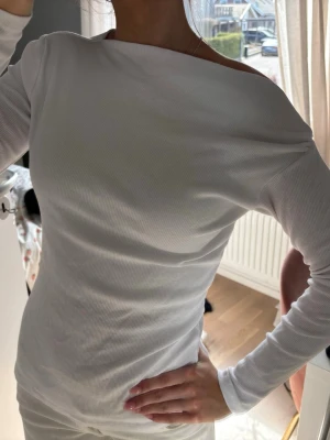 Vit offshoulder topp - Säljer en vit ribbad topp med offshoulder-design och lång ärm. Toppen har en slim passform och är tillverkad i ett mjukt, stretchigt material som sitter snyggt på kroppen. Perfekt för dig som gillar stilrena och trendiga plagg.