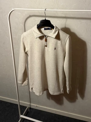 Beige långärmad polotopp från Polo Ralph Lauren - Säljer en beige långärmad polotopp från Polo Ralph Lauren med klassisk krage och half zip-dragkedja framtill. Diskret logga broderad på bröstet. Materialet är mjukt och ribbat, perfekt för en stilren look.