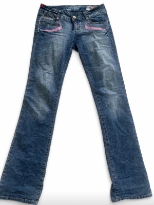 Blå bootcut jeans med rosa brodyr - Snygga blå bootcut jeans med rosa broderade detaljer på fickorna fram och bak. Jeansen har låg midja och klassisk femficksdesign. Tillverkade i denim med lätt slitning för en cool look.