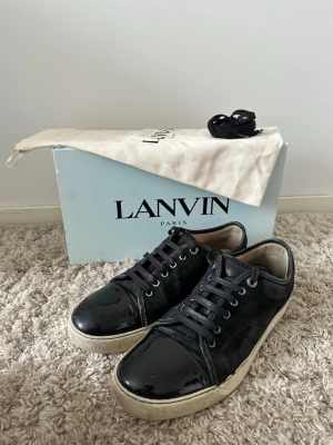 Svarta Lanvin sneakers i mocka och lack - Snygga svarta sneakers från Lanvin med ovandel i mocka och tåparti i blankt lackläder. Klassisk låg modell med vita sulor och svarta skosnören. Kommer med originalkartong och dustbag. Perfekta för dig som gillar exklusiva och stilrena sneakers. Allt på bilden ingår 