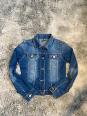 Klassisk blå jeansjacka - Snygg blå jeansjacka med klassisk passform och bröstfickor med knapp. Jackan har långa ärmar, knappar framtill och detaljer i guld. Perfekt att slänga över en hoodie eller t-shirt för en cool look.Den är i storlek 152 