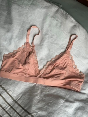 Rosa spetsbralette med trekantskupor - Söt ljusrosa bralette med mjuka trekantskupor och dekorativ spets längs kanterna. Justerbara axelband och bred resår under bysten för extra komfort. Perfekt för dig som gillar en enkel men ändå feminin stil.
