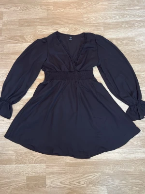 Svart kort omlottklänning med spets - Svart kort klänning från Gina Tricot med omlott-skärning och v-ringning med spets. Klänningen har långa ärmar med resår vid handlederna och markerad midja. I storlek M