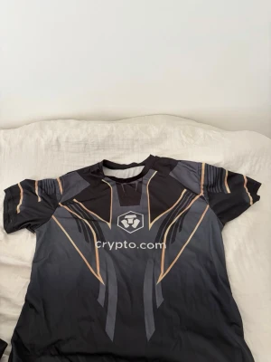 Svart sporttröja Crypto.com Makhachev - Snygg svart sporttröja med guld- och gråa detaljer, Crypto.com-logga på bröstet och namnet Makhachev på ryggen. Tillverkad i ett lätt och snabbtorkande material, perfekt för träning eller e-sport. Modern design med korta ärmar och rund hals.