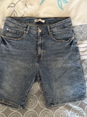Blå jeansshorts från Zara - Säljer ett par klassiska blå jeansshorts från Zara med fem fickor och knappgylf. Shortsen har en avslappnad passform och är tillverkade i slitstark bomull. Perfekta för varma dagar och enkla att matcha med olika stilar.