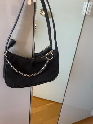 Svart axelväska med kedjedetalj - Trendig svart axelväska  från Åhléns med silverfärgad kedja framtill och dragkedja upptill. Väskan har en mjuk, rundad form. Perfekt för att bära det viktigaste och ger en cool touch till din outfit.